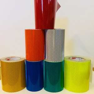 May include: A stack of seven rolls of reflective tape in various colors, including red, orange, silver, yellow, gold, blue, and green. The tape is designed for safety and visibility.