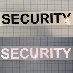 May include: Two rectangular stickers with the word "SECURITY" printed in a bold, sans-serif font. The top sticker has black text on a white background, while the bottom sticker has light pink text.