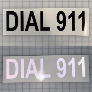 May include: Two rectangular signs with the text "DIAL 911" in black and white. The top sign has black text on a white background. The bottom sign has white reflective text on a gray background.