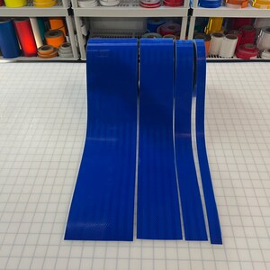 Puede incluir: Cuatro rollos de cinta reflectante azul de diferentes anchos. La cinta est&aacute; sobre una superficie de rejilla blanca.