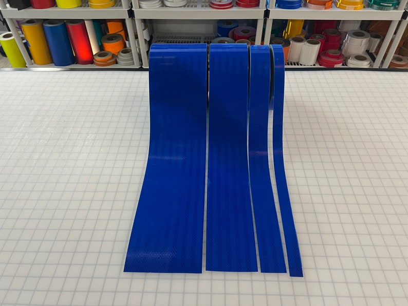 Puede incluir: Cuatro rollos de cinta reflectante azul de diferentes anchos. La cinta est&aacute; sobre una superficie de rejilla blanca.