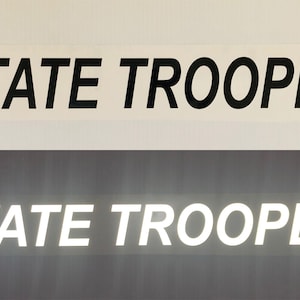 May include: Two images of a reflective sticker with the text "STATE TROOPER" in black lettering on a white background. The top image shows the sticker on a white surface, while the bottom image shows the sticker on a dark gray surface.
