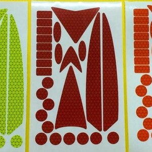 May include: Three sheets of reflective stickers in yellow, red, and orange. Each sheet contains a variety of shapes and sizes of stickers, including circles, triangles, and rectangles. The stickers are designed to be used on bicycles, helmets, and other items to increase visibility.