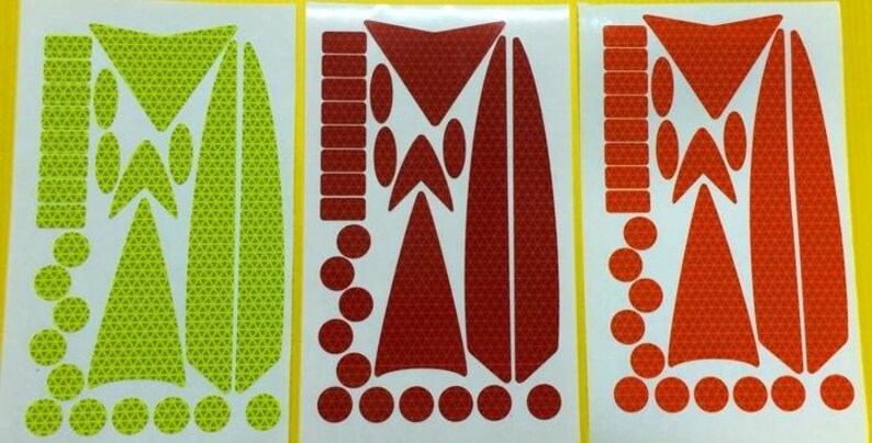 May include: Three sheets of reflective stickers in yellow, red, and orange. Each sheet contains a variety of shapes and sizes of stickers, including circles, triangles, and rectangles. The stickers are designed to be used on bicycles, helmets, and other items to increase visibility.