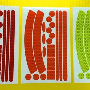 May include: Three sheets of reflective bike stickers. The stickers are in red, orange, and green. Each sheet has a variety of shapes and sizes of stickers, including circles, rectangles, and curved lines.