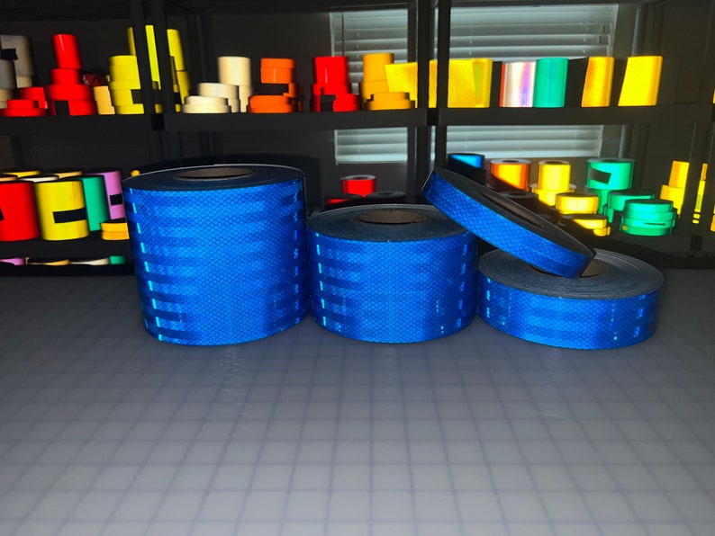 Puede incluir: Cuatro rollos de cinta reflectante azul, con un patr&oacute;n de diamante, est&aacute;n apilados sobre una superficie gris. La cinta se utiliza para la seguridad y la visibilidad.