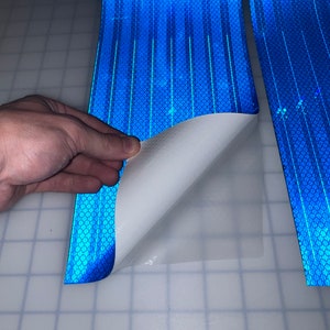 Puede incluir: Un rollo de cinta reflectante azul con un respaldo adhesivo blanco. La cinta se est&aacute; despegando para revelar el adhesivo.