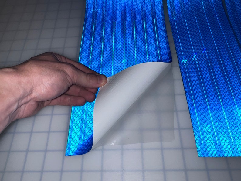 Puede incluir: Un rollo de cinta reflectante azul con un respaldo adhesivo blanco. La cinta se est&aacute; despegando para revelar el adhesivo.