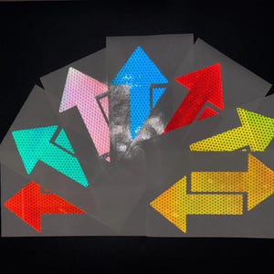 May include: Assortment of reflective arrow stickers in various colors, including blue, pink, red, yellow, and green. The stickers are on a white background and have a grid pattern.