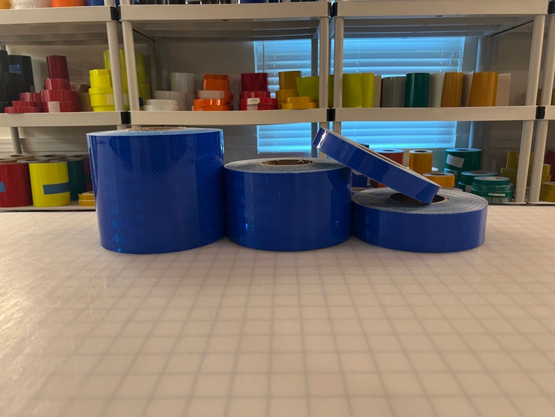 Puede incluir: Cuatro rollos de cinta reflectante azul. La cinta est&aacute; sobre una superficie de rejilla blanca.