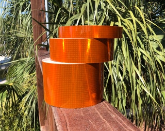 May include: Three rolls of orange reflective tape. The tape is diamond shaped and has a reflective surface.
