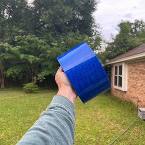 Puede incluir: Un rollo de cinta reflectante azul con un patr&oacute;n de diamante. La cinta se sostiene en una mano.