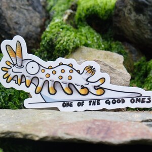 May include: A white and yellow cartoon sea slug sticker with the text "One of the good ones" on a white background. The sticker is on a bed of green moss.