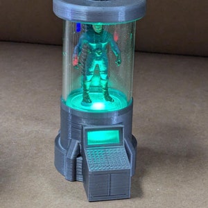 May include: A gray and clear plastic tube with a black hose on top. Inside the tube is a small gray figure with a green light shining from the bottom of the tube.