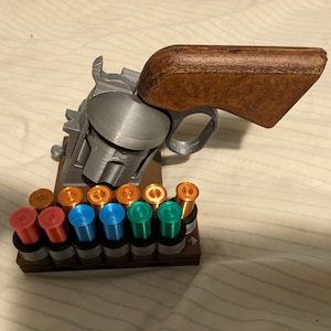 May include: A 3D-printed toy revolver with a brown handle and a silver-coloured body. The revolver is displayed with a rack of colourful bullets in red, orange, blue, and green. The toy gun is a novelty item.
