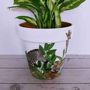 May include: White ceramic planter with a jungle scene featuring a zebra, tiger, and giraffe. The planter is filled with a green leafy plant.