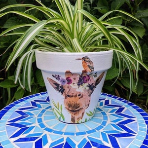 May include: A white ceramic planter featuring a painted giraffe design, adorned with a floral crown and a small bird perched on top. The planter is placed on a blue and white mosaic tile surface.