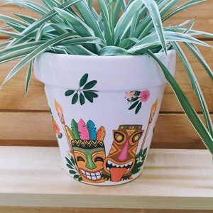 May include: A white ceramic planter with a hand-painted design featuring two tiki masks, green leaves, and pink flowers. A spider plant with long, green and white striped leaves is growing in the planter.
