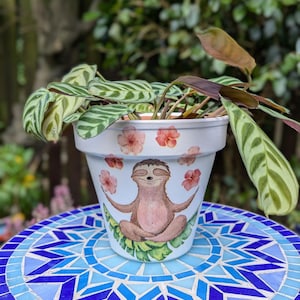 May include: White ceramic planter with a hand-painted design of a sloth meditating on a green leaf. The planter has pink flowers around the sloth and is filled with a green and white striped plant.