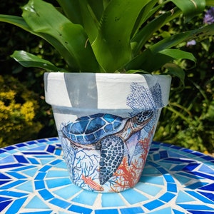 May include: A white ceramic planter featuring a blue and orange sea turtle design. The planter is decorated with coral and seaweed. The planter is sitting on a blue and white mosaic table.