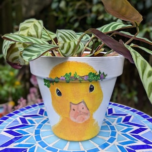 May include: A white ceramic planter with a hand-painted yellow duckling face. The duckling is wearing a purple flower crown. The planter is filled with a green and white striped plant.