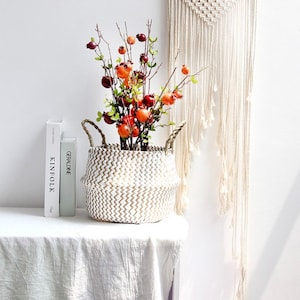 May include: A decorative white woven basket with a zig-zag pattern, filled with faux pomegranate branches. The pomegranates are red and orange, with green leaves. Two books, "KINFOLK" and "GERALDINE", are on the left. A macrame wall hanging is in the background.