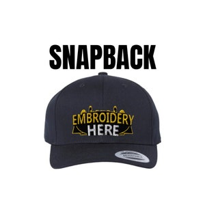 May include: A navy blue snapback hat with the text "Embroidery Here" in gold and white lettering.