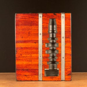 以下が含まれることがあります： 銀色の金属製のストリップとネジが付いた木製のディスプレイケースに、黒い金属製のカムシャフトが取り付けられています。木は赤褐色です。