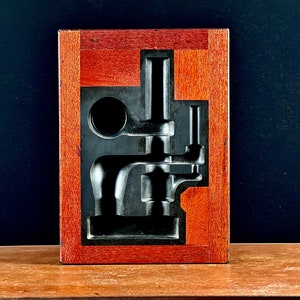 以下が含まれることがあります： 木製の箱の中に、濃い灰色の金属製の型が入っています。型は、複数の部品を持つ複雑な機械装置の形をしています。