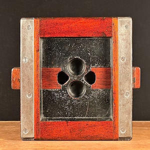 May include: A vintage wooden camera plate with a metal frame and three circular holes. The plate is a dark reddish brown color and the frame is silver. The plate has a black surface with a worn look.