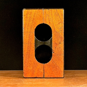 以下が含まれることがあります： 2つの円形の切り抜きのある木製のブロック。ブロックは濃い茶色に着色されており、前面にグリッドパターンがあります。