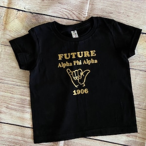 May include: Black t-shirt with gold lettering that reads "FUTURE Alpha Phi Alpha 1906" and a gold hand gesture.