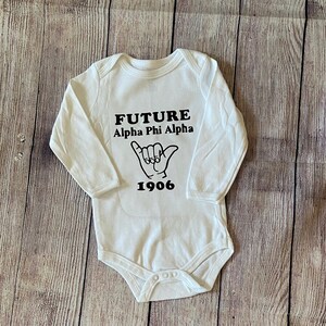 May include: White long-sleeved baby onesie with black text that reads "FUTURE Alpha Phi Alpha 1906" and a black hand gesture graphic.
