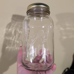 Puede incluir: Un tarro de cristal transparente con tapa dorada. El tarro tiene un diseño blanco de batidora y burbujas, con el nombre "Ruth" en fuente cursiva. El tarro se utiliza probablemente para el almacenamiento en la cocina.