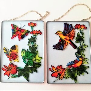 May include: Two square glass panels with metal frames and hanging chains. Each panel features a painted scene of butterflies and birds with flowers and green leaves. The butterflies are orange, yellow, and black. The birds are yellow, orange, red, and blue. The flowers are red and orange.