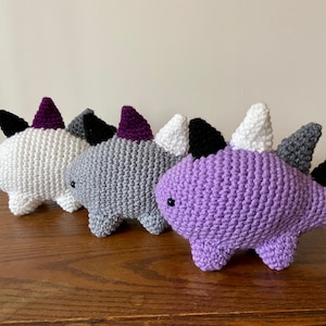 May include: Four crocheted dinosaur plushies in different colors: white, gray, purple, and black. The dinosaurs have spiky backs and are arranged in a row.