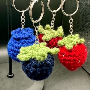 May include: Four crocheted fruit keychains: two blue blueberries and two red strawberries with green leaves. The keychains are attached to silver key rings.