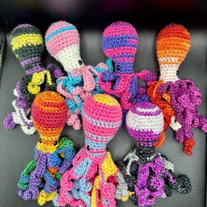 May include: A collection of seven colorful, crocheted octopus toys. Each octopus features a striped head in various colors like pink, orange, and purple, with contrasting tentacles. The toys are handmade and appear soft and plush.