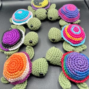 May include: A collection of handmade crochet turtles in various colors. The turtle shells feature vibrant patterns in shades of blue, pink, purple, orange, and yellow. The turtles have green bodies and black eyes, creating a whimsical and colorful display.