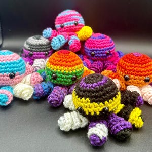 May include: A collection of handmade crochet octopus toys in various colors. The octopuses feature a variety of vibrant hues, including pink, blue, purple, orange, yellow, and rainbow patterns. Each octopus has black button eyes and a rounded head.