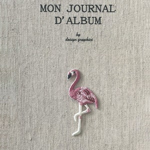 May include: A journal with the text "MON JOURNAL D'ALBUM" and a flamingo patch. The journal cover is a light beige fabric. The flamingo is pink with white legs and a black beak. The text "by design graphics" is also visible.