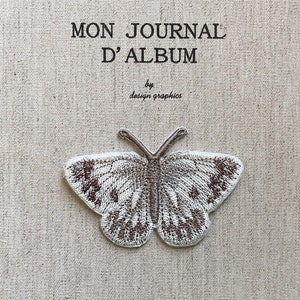 May include: Embroidered butterfly patch in brown and white on a beige fabric. The French text "MON JOURNAL D'ALBUM" is above the butterfly, with "by design graphics" below. The patch is a decorative element, suitable for crafts or embellishment.