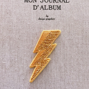 Może przedstawiać: Złota, haftowana naszywka w kształcie błyskawicy na teksturowanej, jasnoszarej tkaninie. Tekst "MON JOURNAL D'ALBUM" znajduje się nad naszywką, a pod nią "by design graphics". Naszywka jest elementem dekoracyjnym.