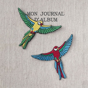 Può includere: Due toppe ricamate di pappagalli in volo. Uno è giallo, verde e blu, l'altro rosso, blu e verde. Le toppe sono su uno sfondo di tessuto beige. Il testo "MON JOURNAL D'ALBUM" è visibile.