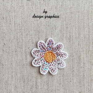 Puede incluir: Una cubierta de diario de tela beige con el texto "MON JOURNAL D'ALBUM" y "by design graphics" impreso en negro. Un parche de margarita brillante con un contorno blanco y un centro naranja está adherido a la cubierta.