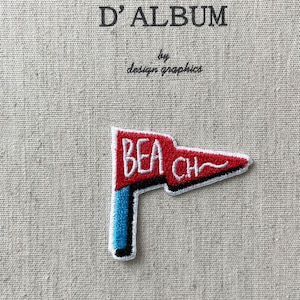 Può includere: Una toppa ricamata rossa e bianca con la scritta "BEACH". Ha la forma di una bandiera con un'asta blu. Lo sfondo è un tessuto beige con il testo "MON JOURNAL D'ALBUM" e "design graphics".