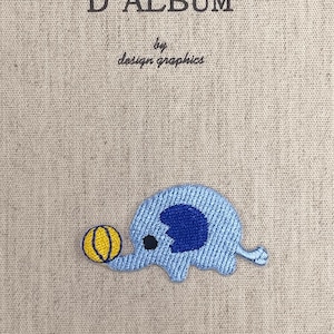 May include: A beige fabric-covered journal with the text MON JOURNAL D'ALBUM and by design graphics printed in dark gray. An embroidered patch of a light blue elephant with a dark blue ear playing with a yellow ball is centered below.