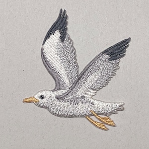 May include: Embroidered patch of a seagull in flight. The bird features gray and white plumage with black wingtips, a yellow beak, and yellow feet. The patch is designed for application on clothing or accessories.