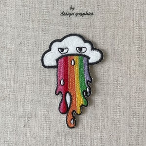 May include: Embroidered patch of a white cloud with a black outline and a grumpy face, vomiting a rainbow. The patch is on a beige fabric background. Text above reads "MON JOURNAL D'ALBUM" and "by design graphics".