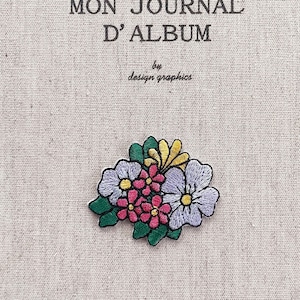 May include: A beige journal cover with the text "MON JOURNAL D'ALBUM" and "by design graphics" in black. A colorful embroidered floral patch with pink, purple, yellow, and green accents is affixed to the cover.